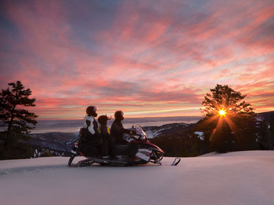Three people on a snowmobile looking out to the setting sun