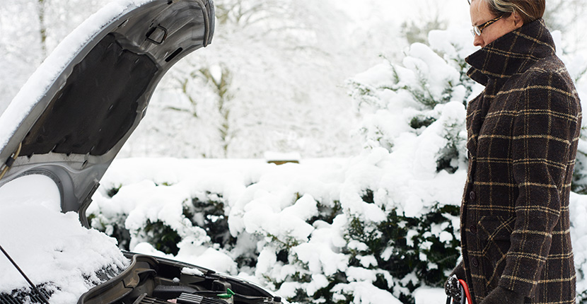a person holding cables looking at a car covered in snow with an opened hood.